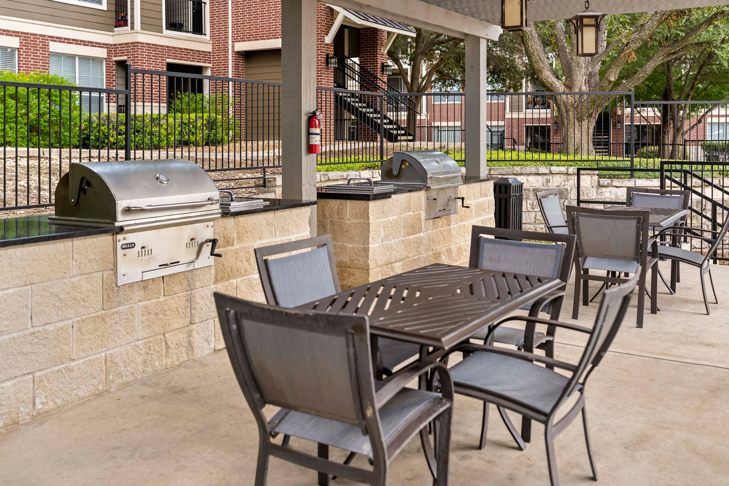 Covered outdoor grilling pavilion with dining tables, stainless grill stations, lantern lighting, and landscaped apartment courtyard setting