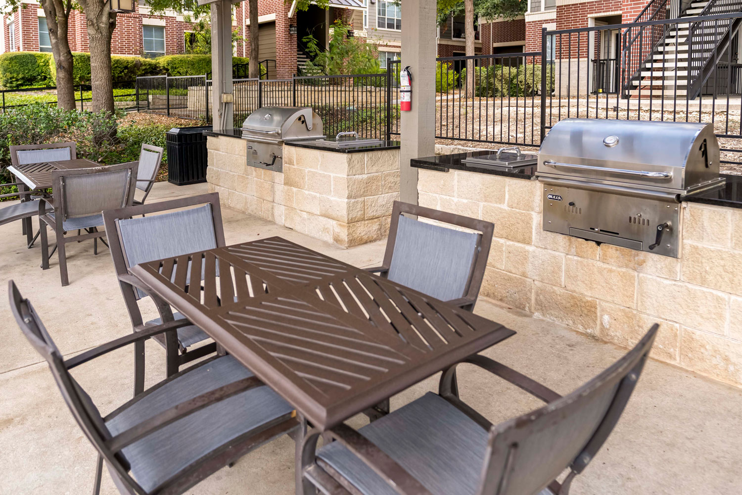 Outdoor dining tables and chairs beside built-in stainless grills under covered pavilion with brick apartment buildings nearby