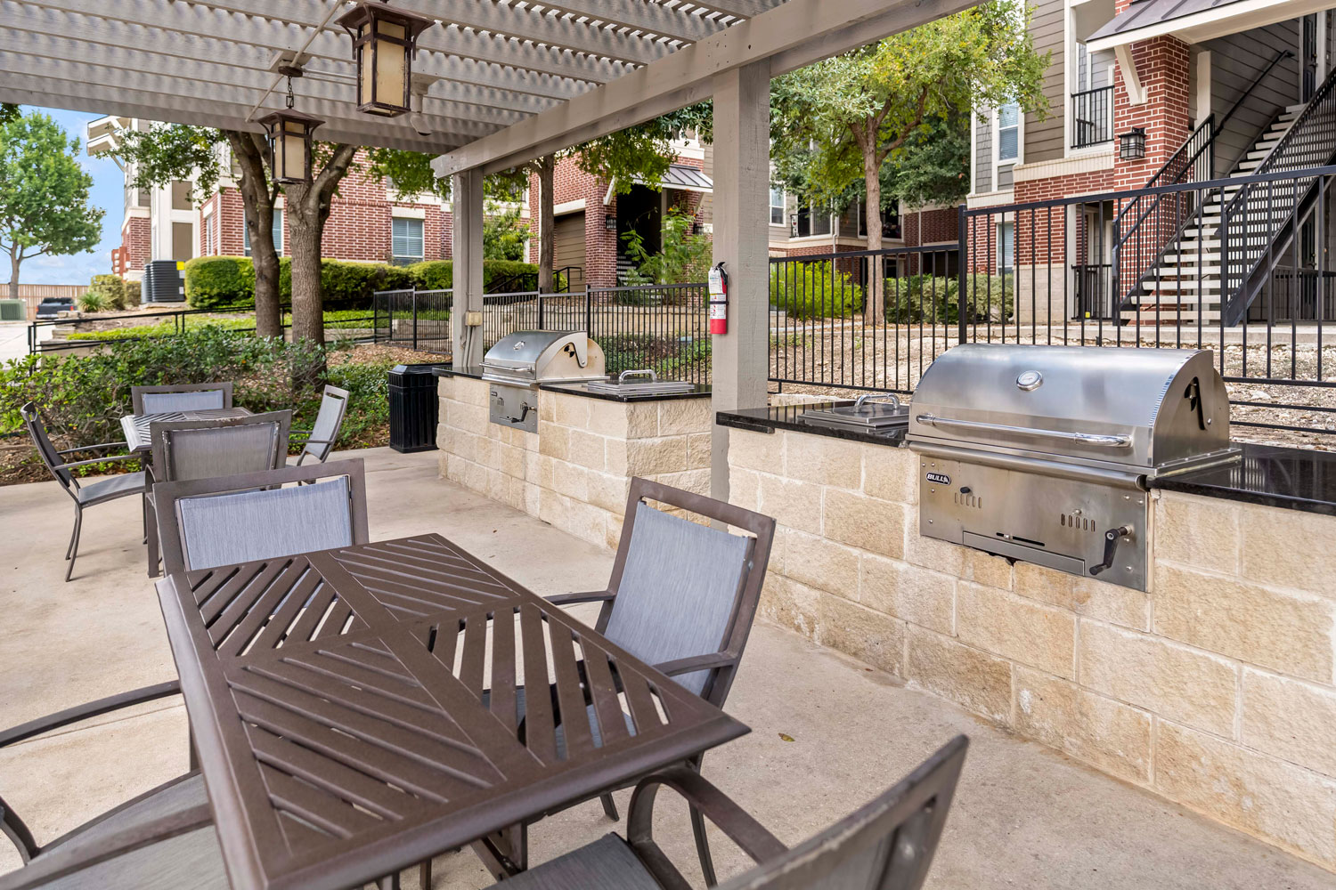 Outdoor grilling pavilion with stainless grills, stone counter workspace, dining tables, lantern lighting, and landscaped courtyard views
