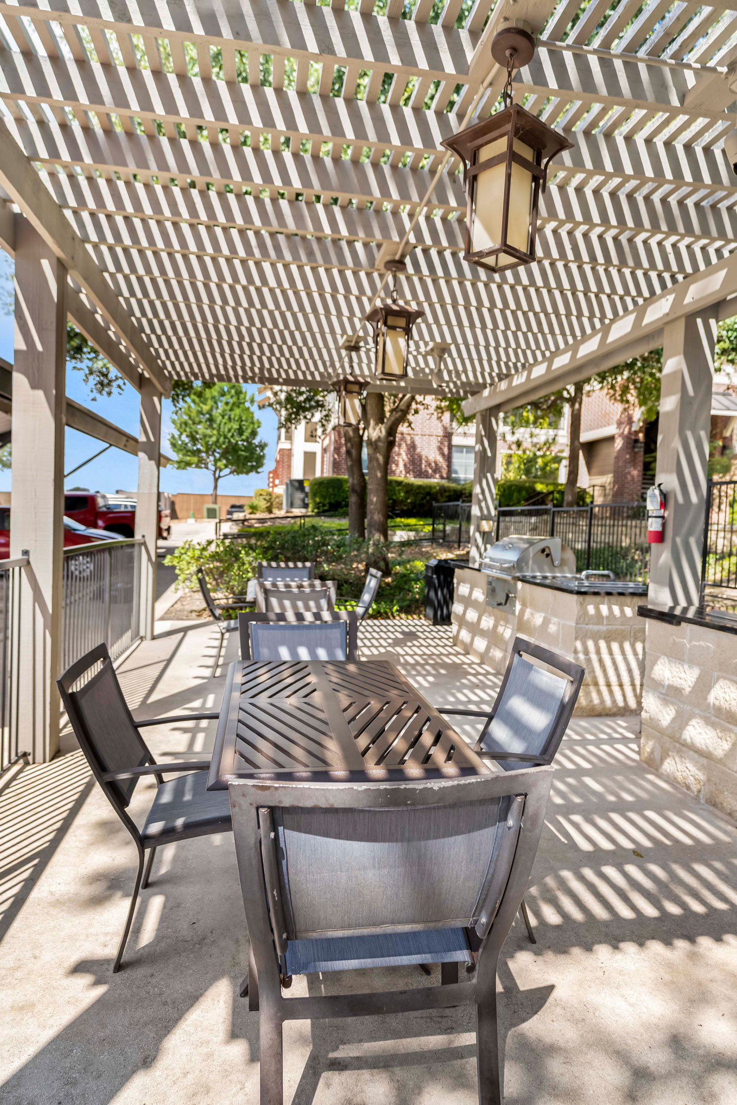 Outdoor grilling pavilion with slatted pergola roof, dining table with chairs, hanging lantern lights, and built-in community grill station
