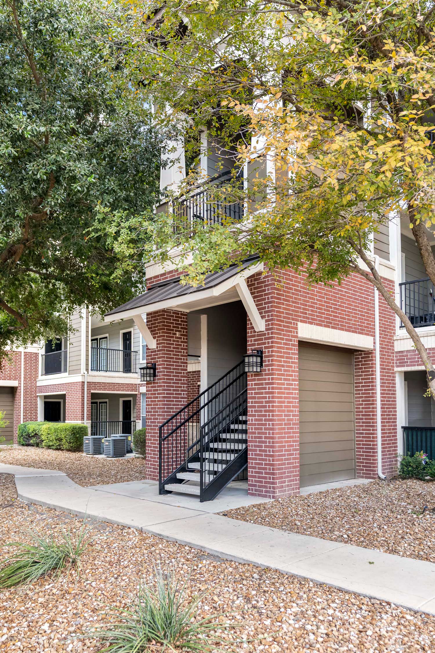 Side view of a brick apartment building with a covered staircase, garage door, and mature trees lining a gravel walkway