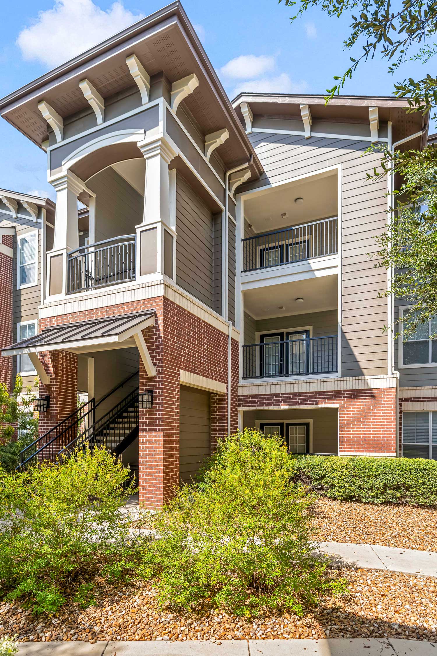 Upward view of a three-story brick and siding apartment building corner with large balconies, decorative columns, and landscaping