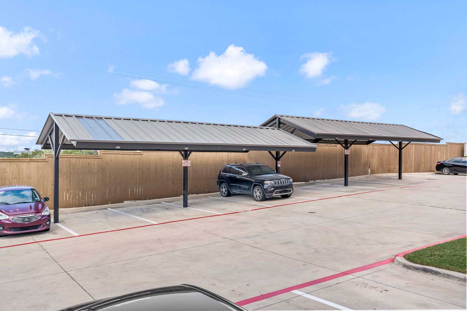 Covered parking area with metal roof carports, designated spaces, perimeter wood fencing, and vehicles parked along paved drive aisle