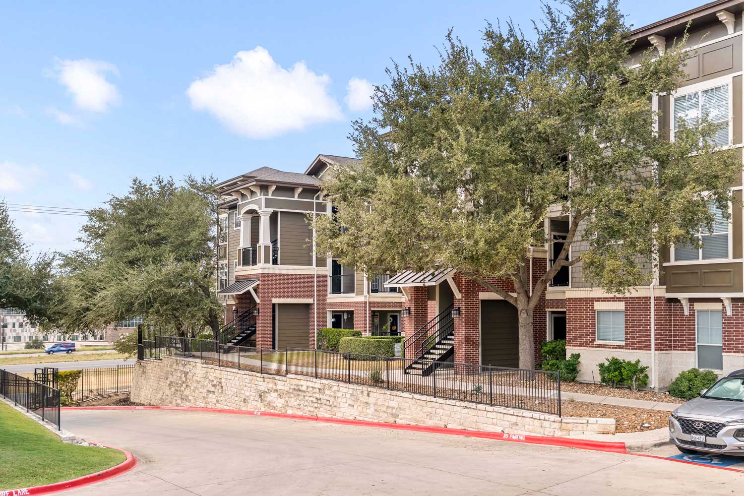Modern apartment community exterior with brick and siding facade, shaded walkways, stair access, and landscaped grounds beside a curved driveway