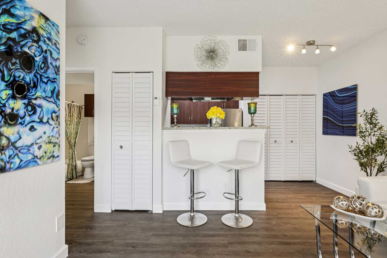 Open concept layout with breakfast bar seating, white louvered doors, wood flooring, and abstract wall art