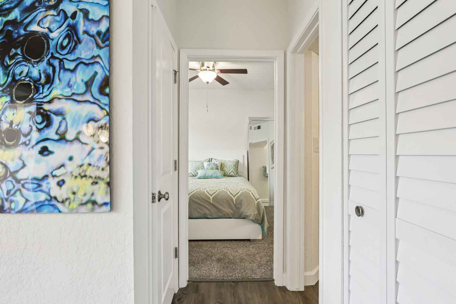 Hallway view featuring abstract wall art, wood-style flooring, louvered closet doors, and bedroom beyond