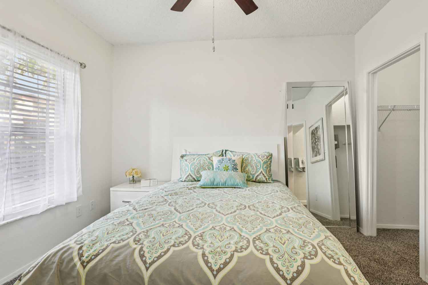 Cozy bedroom with patterned comforter, bedside table, large window, and plush carpet flooring