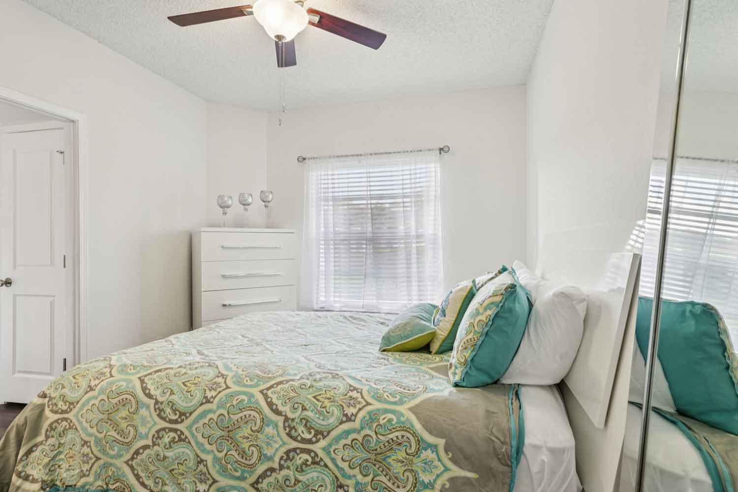Bright bedroom with ceiling fan, mirrored closet doors, window with blinds, and white dresser