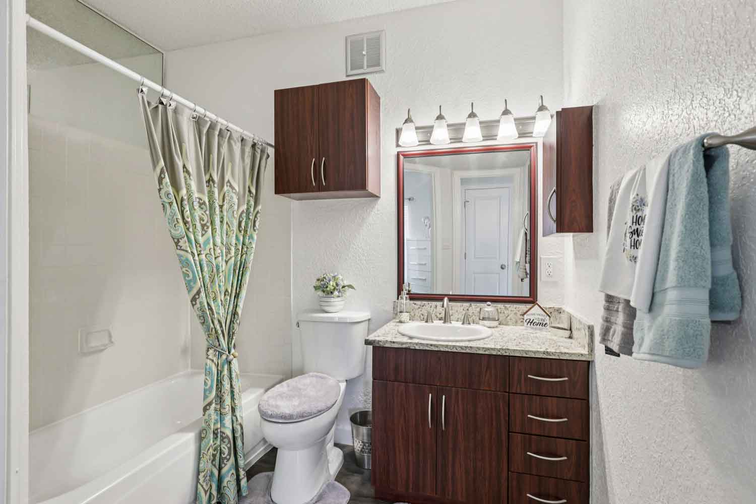 Bathroom with single sink granite vanity, wood cabinetry, overhead lighting, and shower with patterned curtain