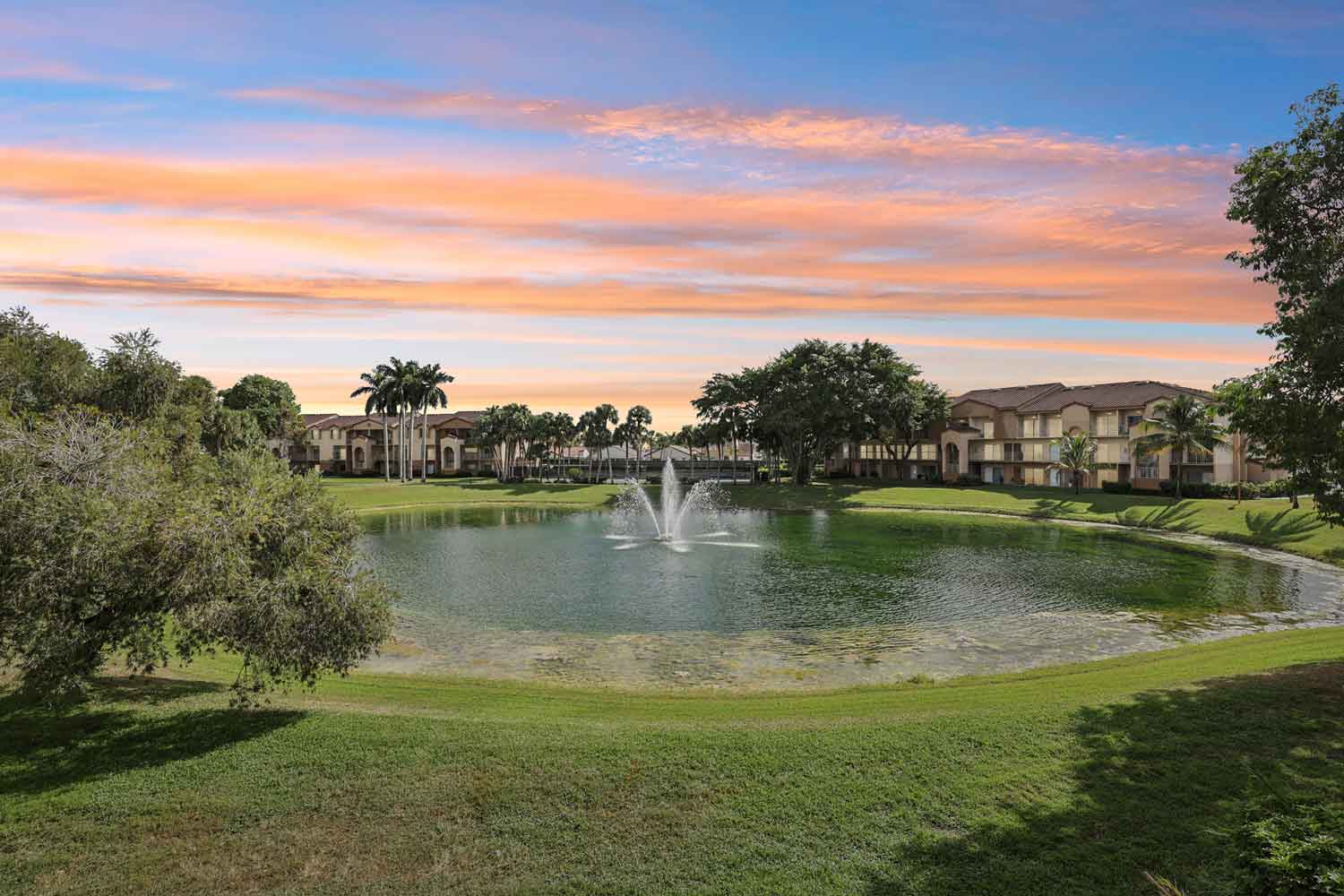 Scenic lake with central fountain, manicured lawns, and apartment buildings at dusk