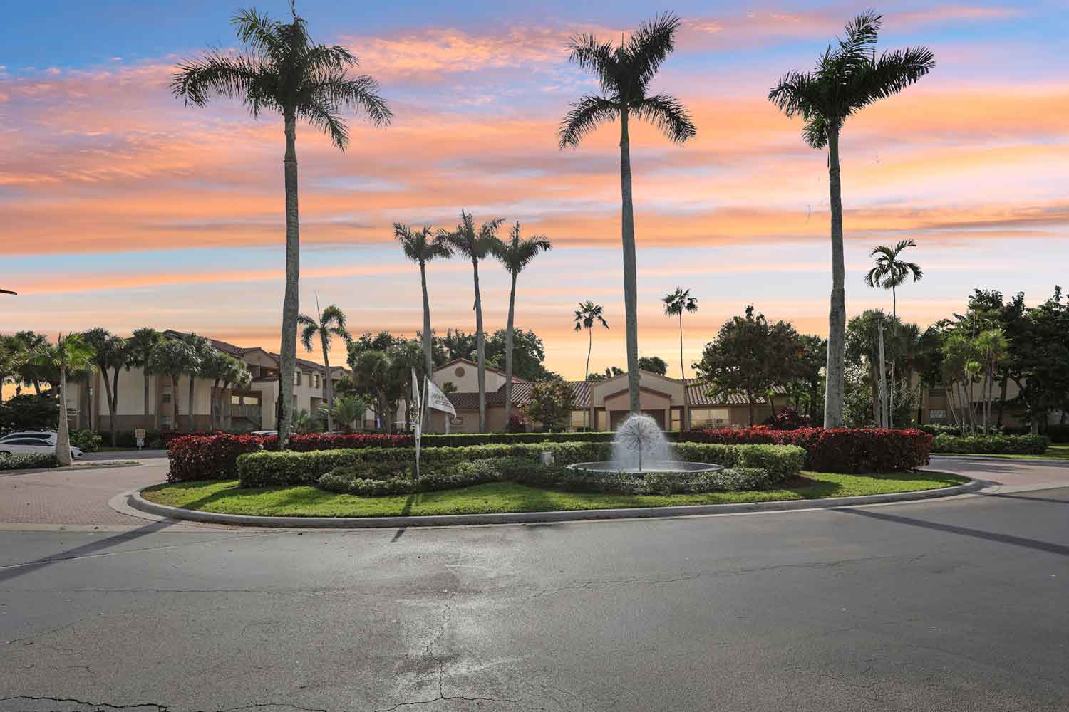 Gated community entrance with palm trees, landscaped roundabout, and fountain at sunset