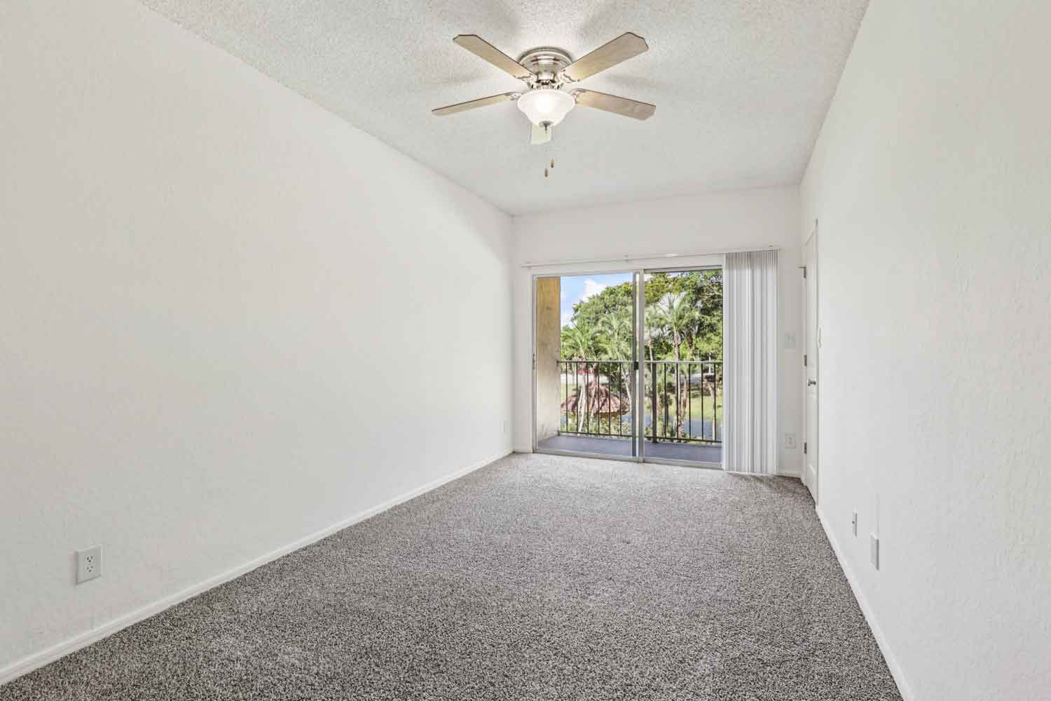 Carpeted bedroom with white walls, ceiling fan, and sliding glass doors opening to a private balcony with palm tree views