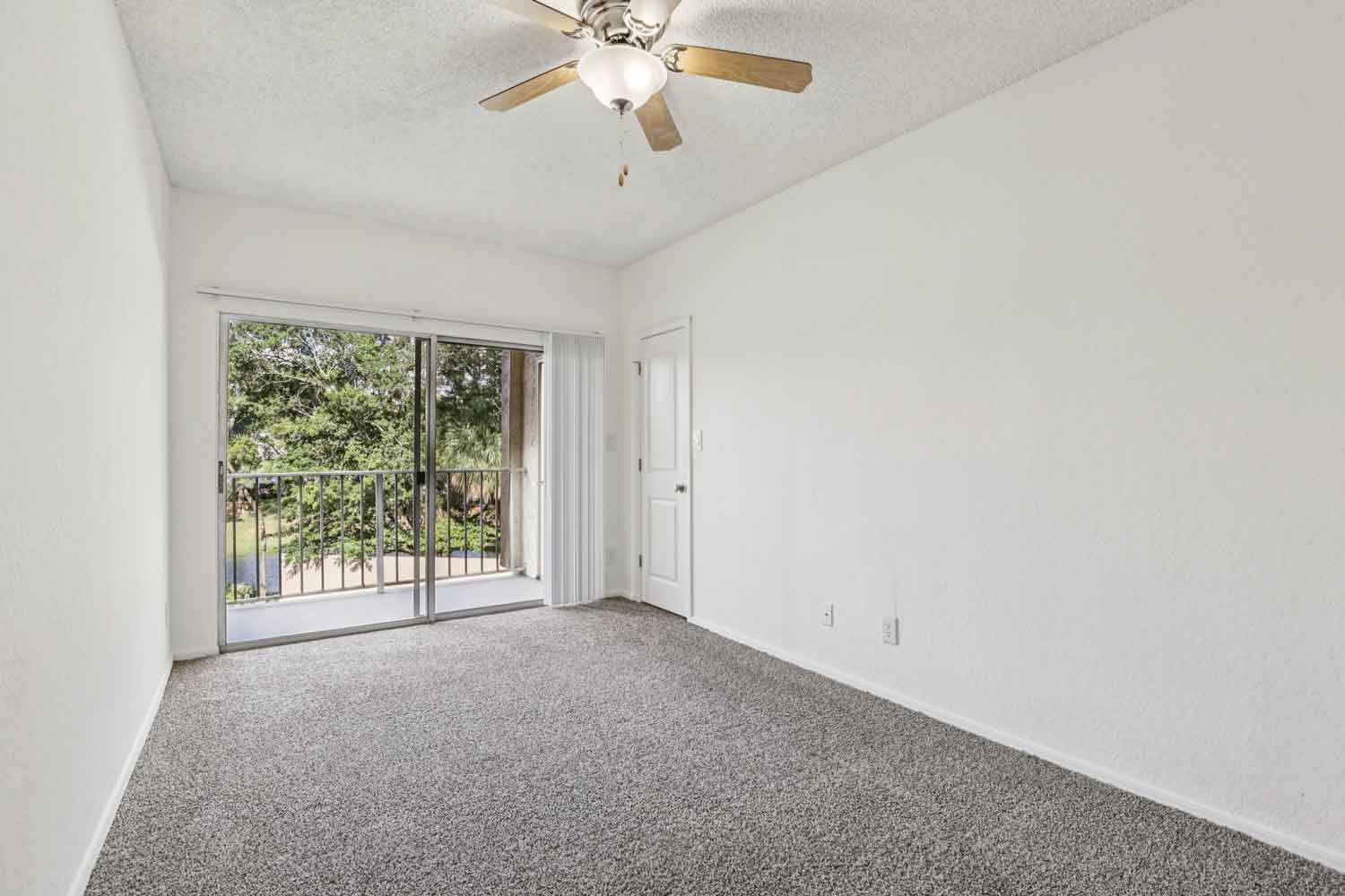 Bright bedroom with ceiling fan, carpet flooring, and sliding glass doors leading to balcony overlooking greenery