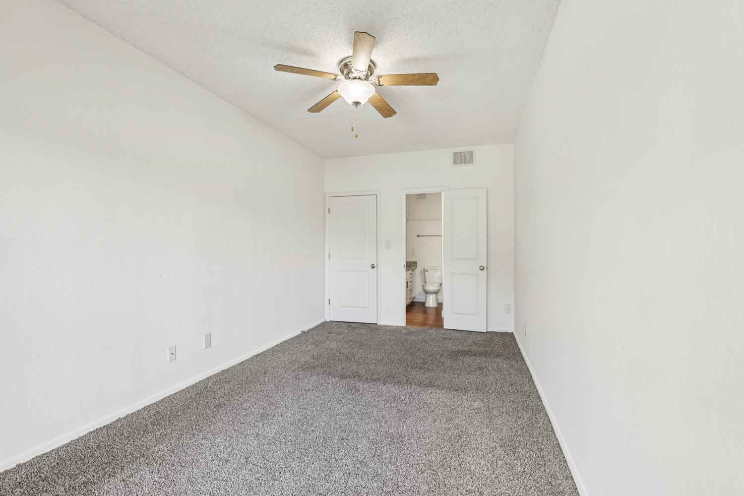 Spacious bedroom with gray carpet, ceiling fan, and open doors revealing attached bathroom