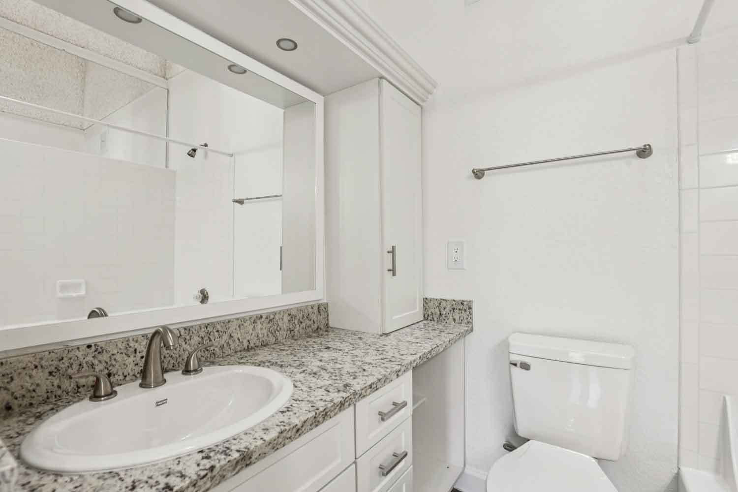 Bathroom vanity with granite countertop, white cabinetry, oval sink, large mirror, and toilet beside tub