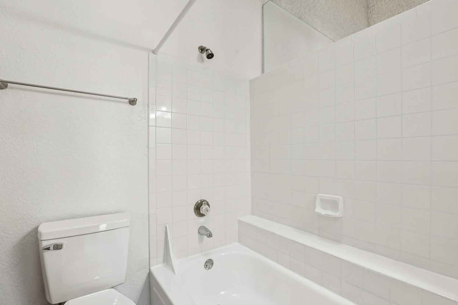 Bathroom with tiled tub and shower combo, chrome fixtures, built in soap shelf, toilet, and towel bar on white wall