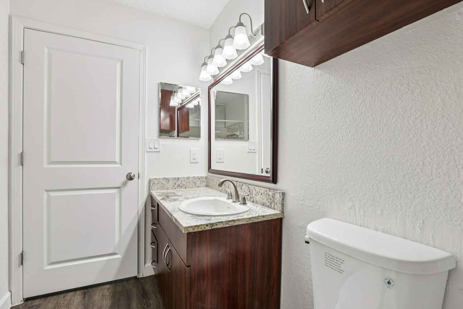 Granite bathroom vanity with oval sink, wood cabinetry, framed mirror, and multi light fixture above countertop