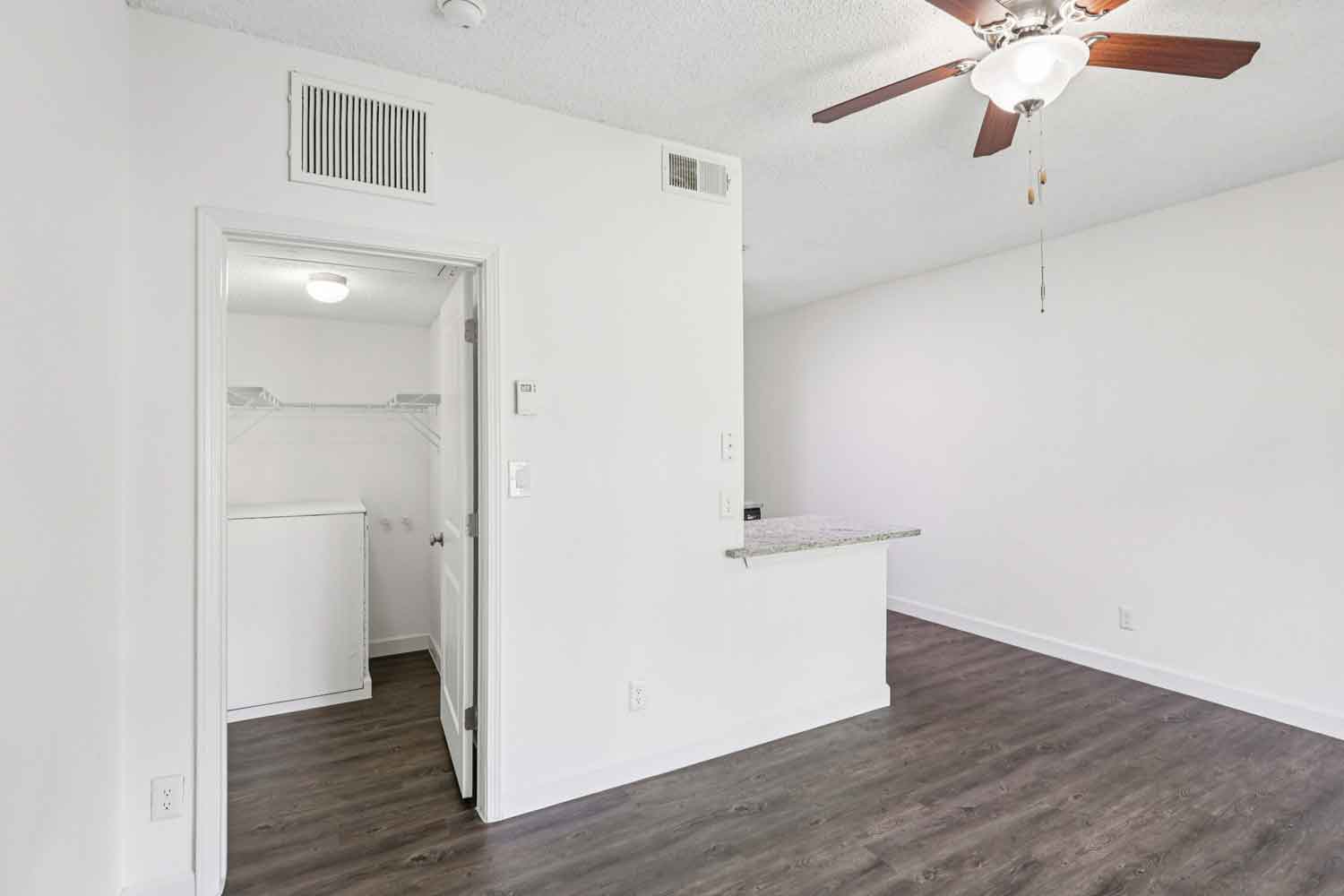 Empty living area with wood style flooring, ceiling fan, breakfast bar, and open doorway to laundry closet