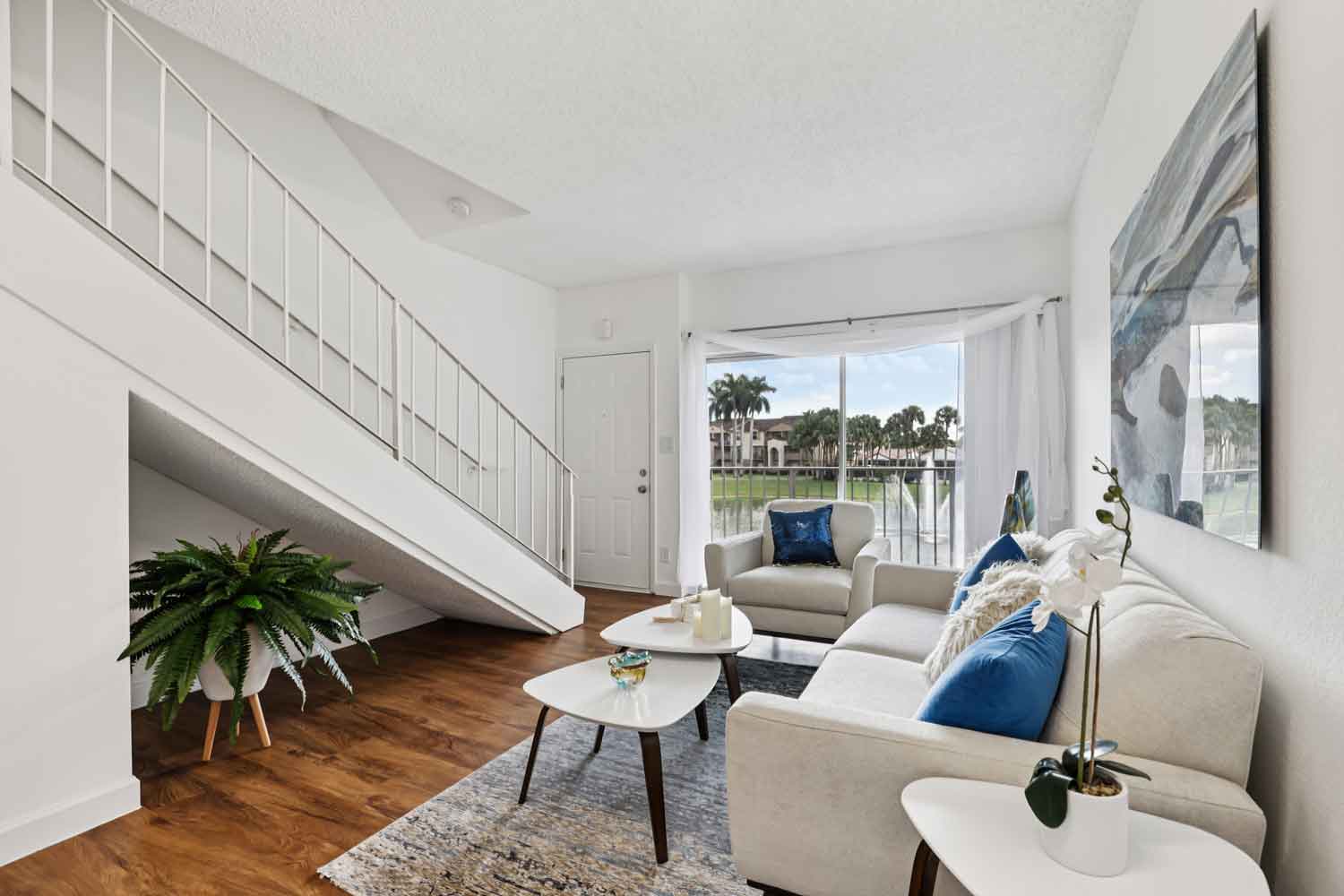 Living room with modern sofa, blue accent pillows, white staircase, coffee table, and large window facing pond