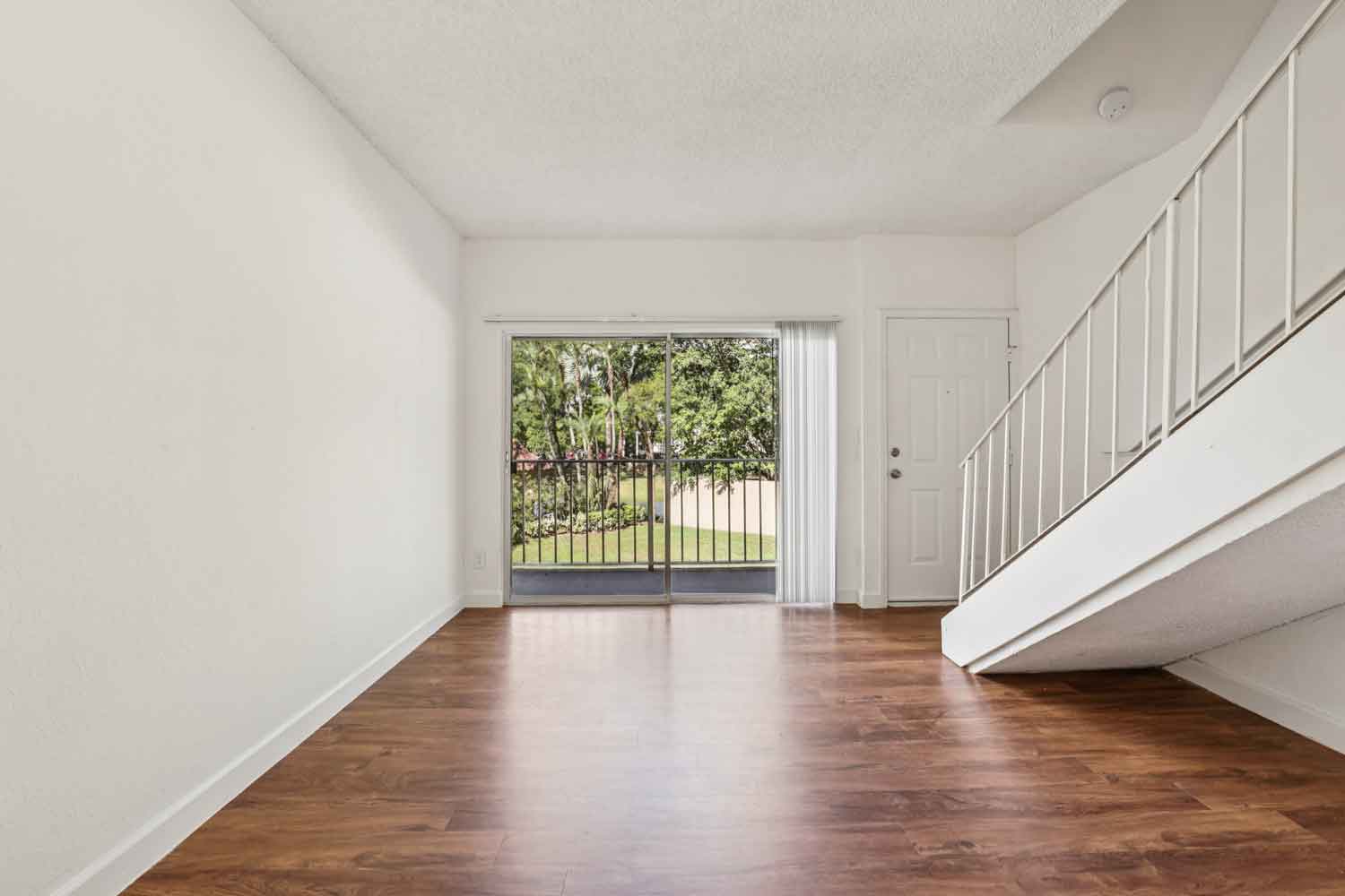 Open living area with wood flooring, staircase with white railing, and sliding glass doors opening to a balcony