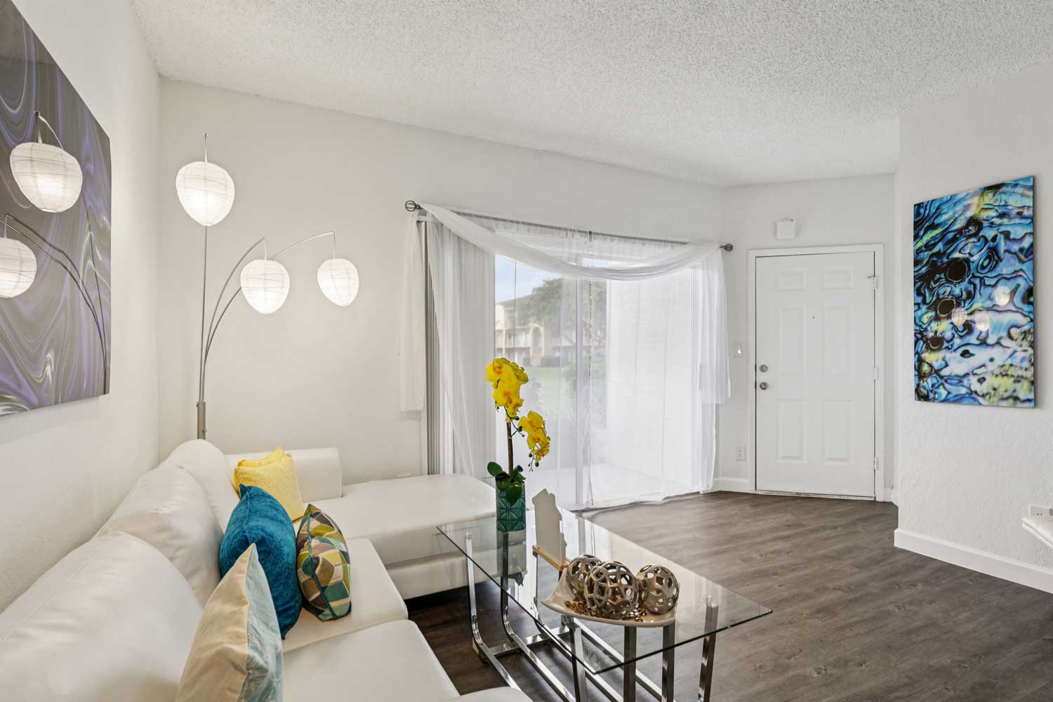 Living room with white sectional sofa, glass coffee table, modern floor lamp, abstract wall art, and sliding glass doors
