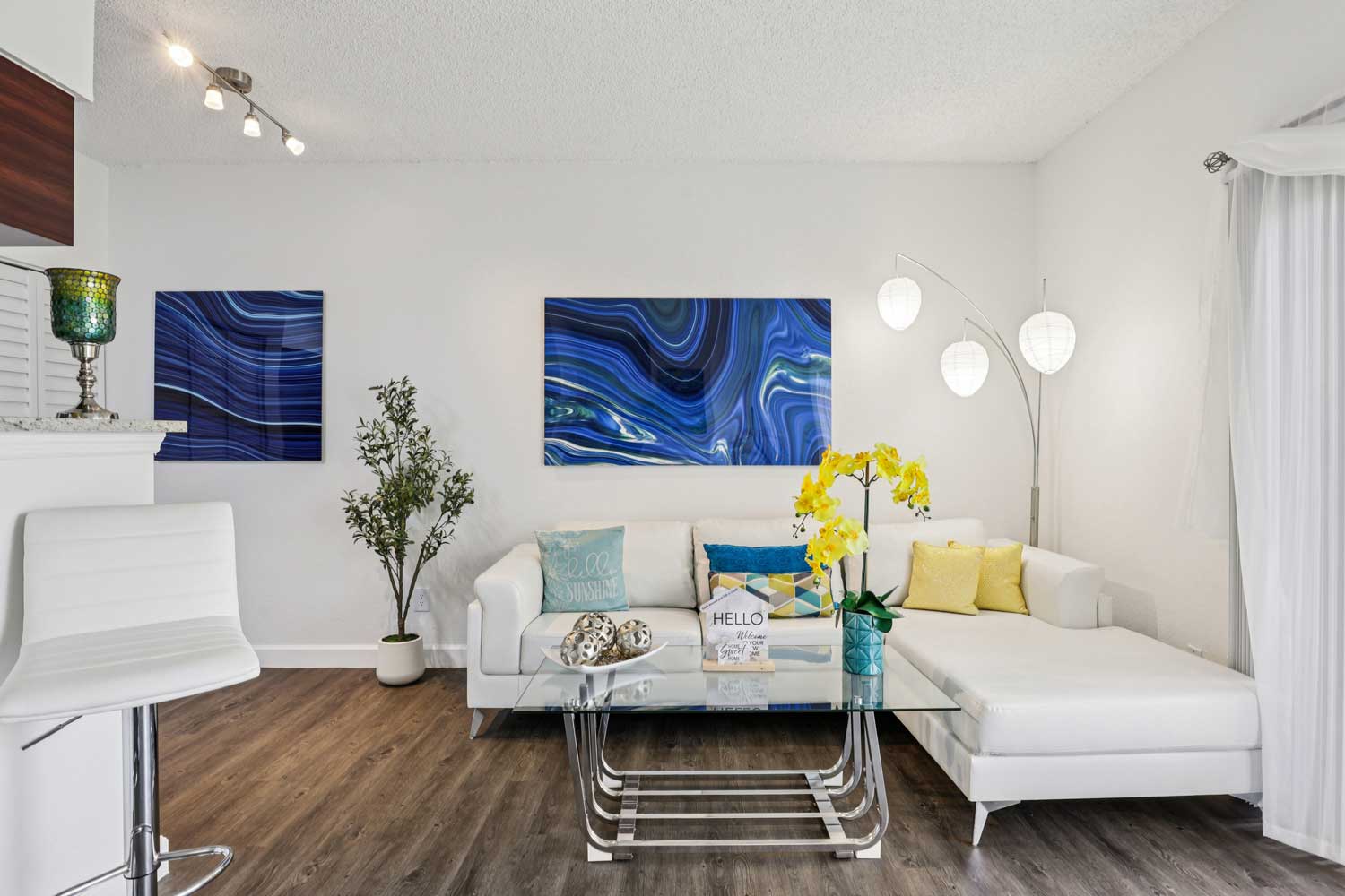 Bright living space with white sectional, blue abstract wall art, glass table, and large window with sheer curtains
