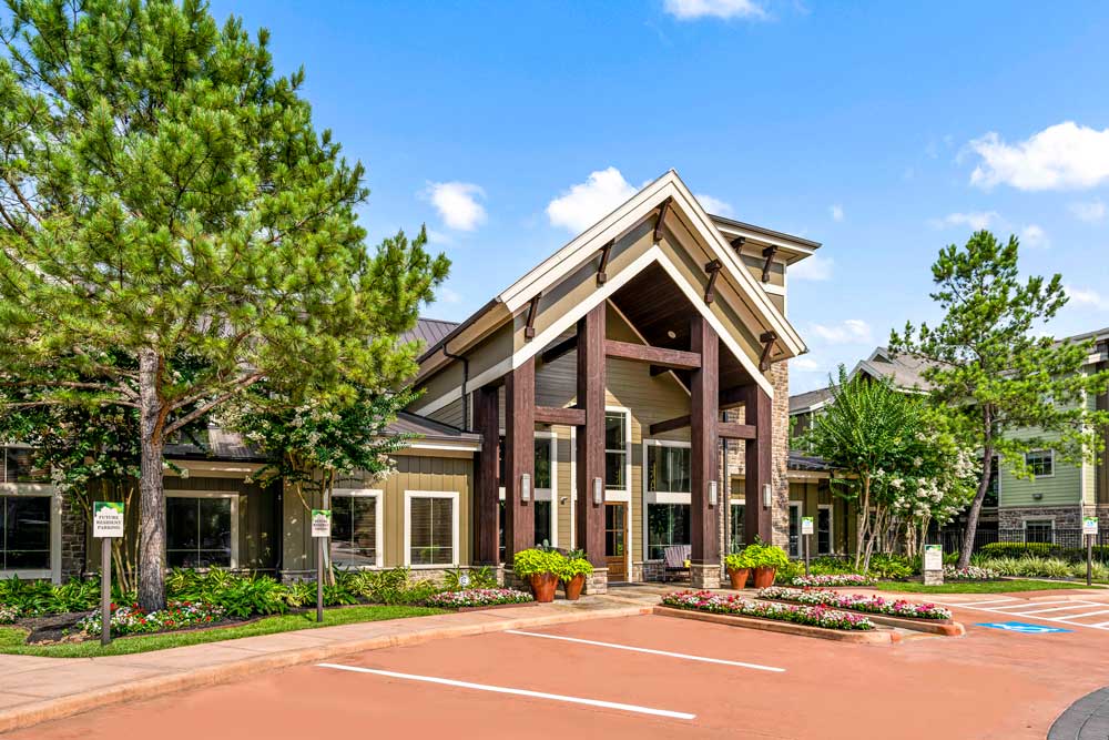 Angled view of The Woodlands Lodge clubhouse with tall trees, garden beds, and front parking area