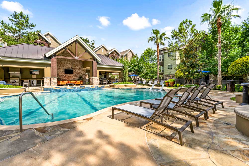 Large outdoor pool with lined-up loungers, palm trees, and a clubhouse featuring stone columns and a TV