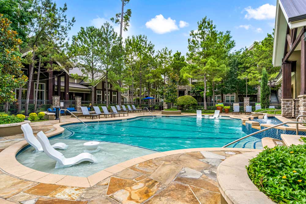 Sunlit pool with built-in loungers, waterfall features, and clubhouse surrounded by pine trees and planters