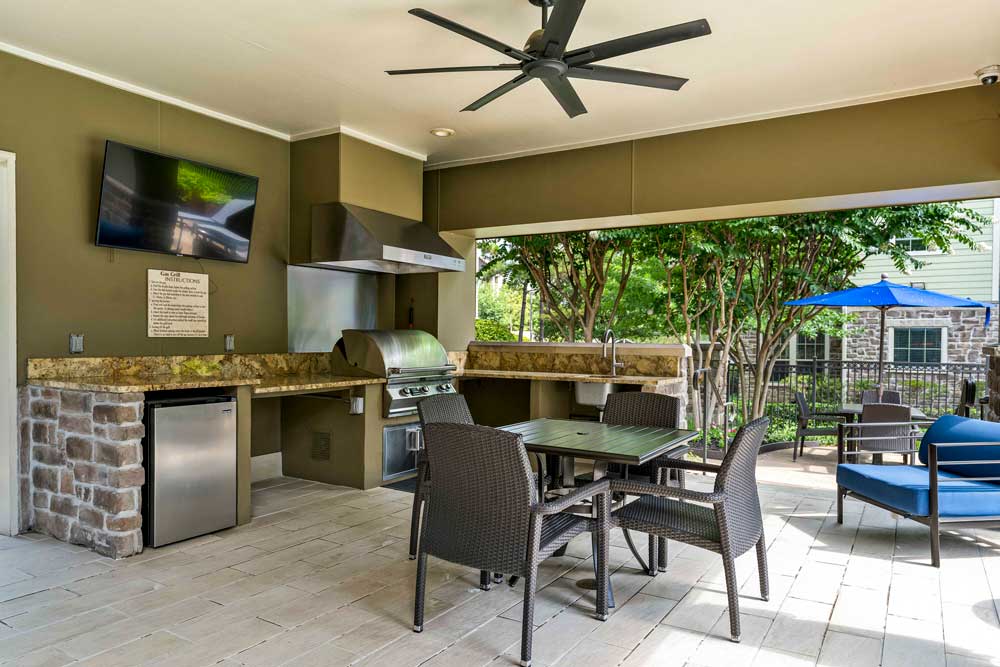 Covered outdoor kitchen with grill, sink, seating area, and wall-mounted TV beside shaded lounge patio and trees