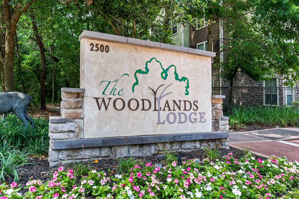 Monument sign for The Woodlands Lodge with address 2500, floral landscaping, and wooded surroundings