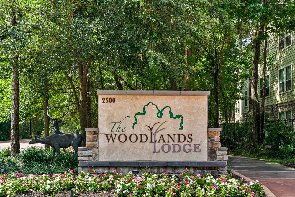 The Woodlands Lodge monument sign with deer sculptures and blooming flowers in a wooded landscape