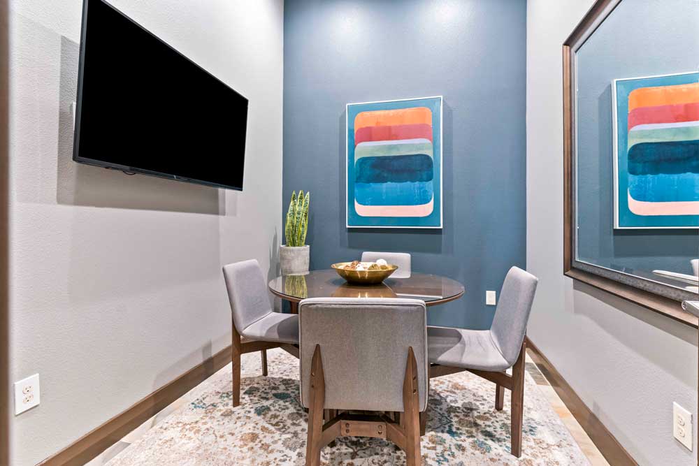 Cozy meeting space with a round table, four gray chairs, wall-mounted TV, and colorful artwork on a blue wall