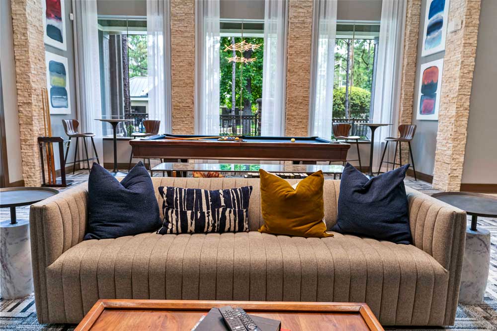 Stylish lounge with a brown sofa, colorful pillows, and a billiards table near tall windows with sheer curtains