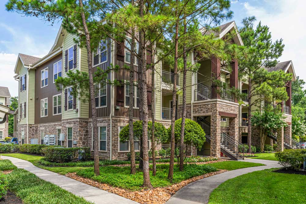 Three-story apartment building with stone and wood exterior, landscaped paths, and surrounding pine trees