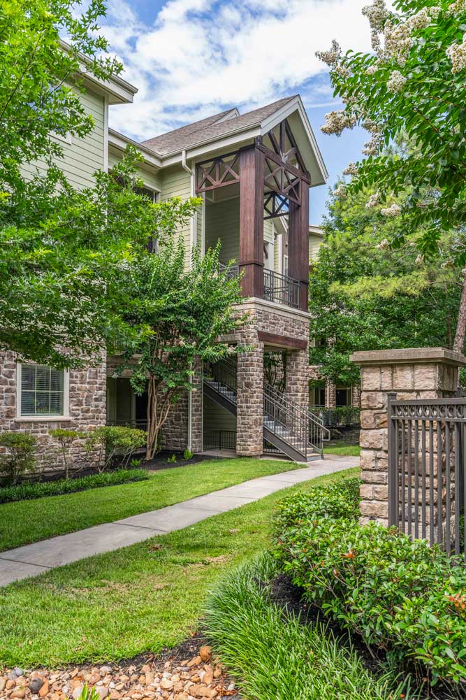 Sidewalk leading to a stone-accented apartment building entrance surrounded by trees and green landscaping
