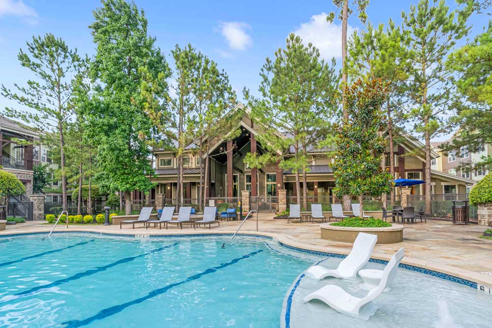 Resort pool with submerged loungers, tall pine trees, and clubhouse in the background under blue skies