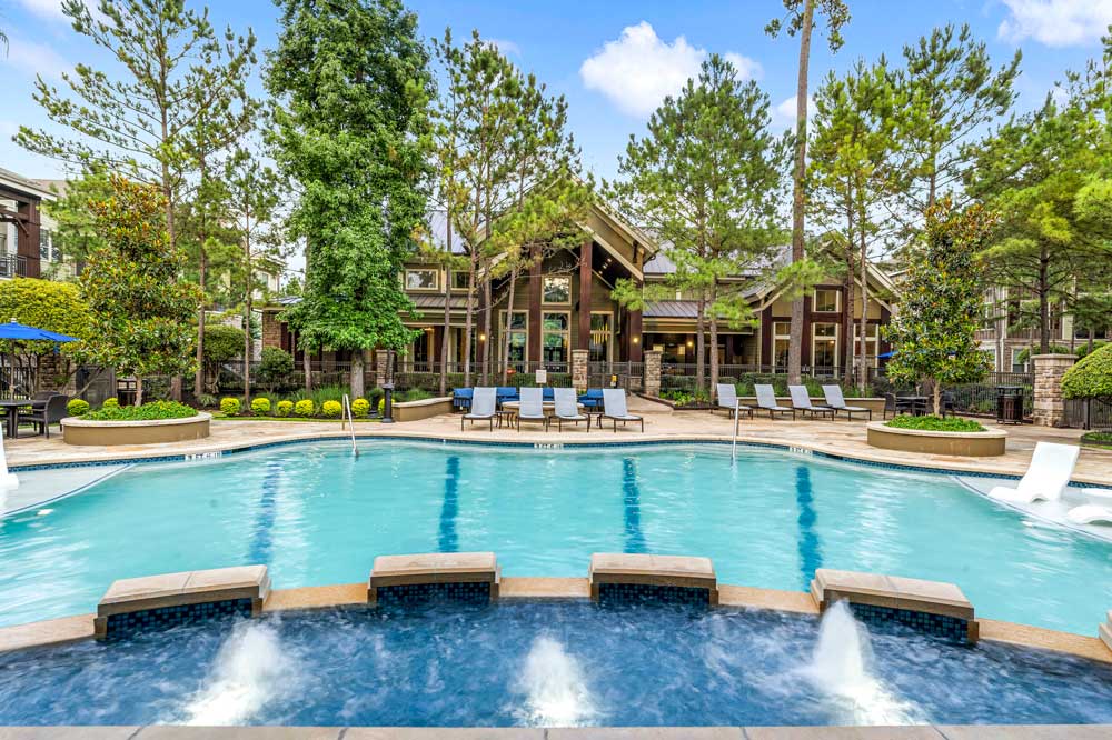 Pool water jets and lounge chairs arranged in front of a grand clubhouse surrounded by lush landscaping
