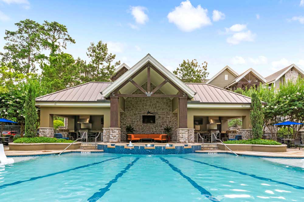 Wide pool with lap lanes and fountains, facing a stone clubhouse under a bright sky with trees and greenery