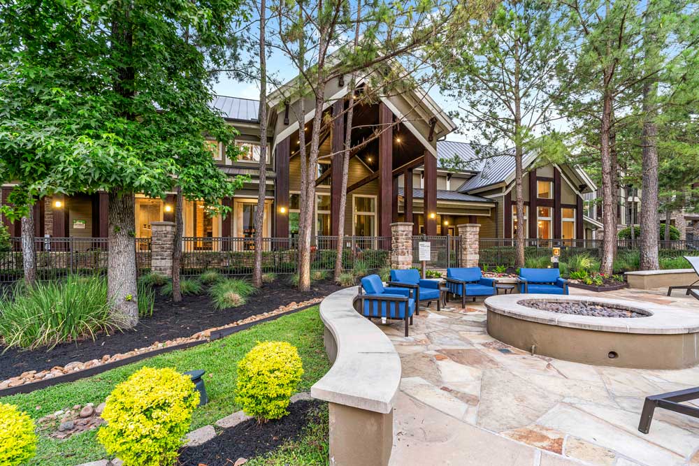 Curved patio seating and fire pit near a glowing clubhouse exterior lined with trees and shrubs