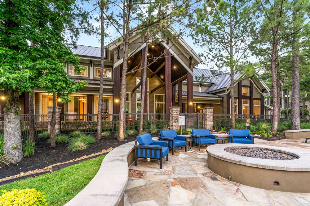 Fire pit area with cushioned chairs in front of a warmly lit clubhouse and tall pine trees
