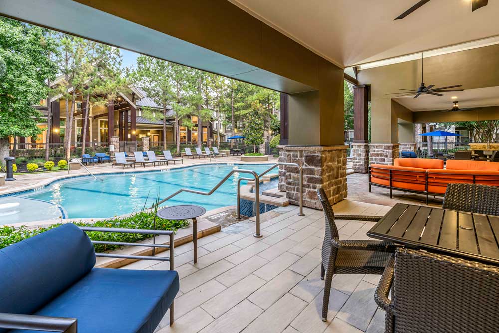 Covered patio with orange and blue seating facing a pool lined with loungers and surrounded by pine trees