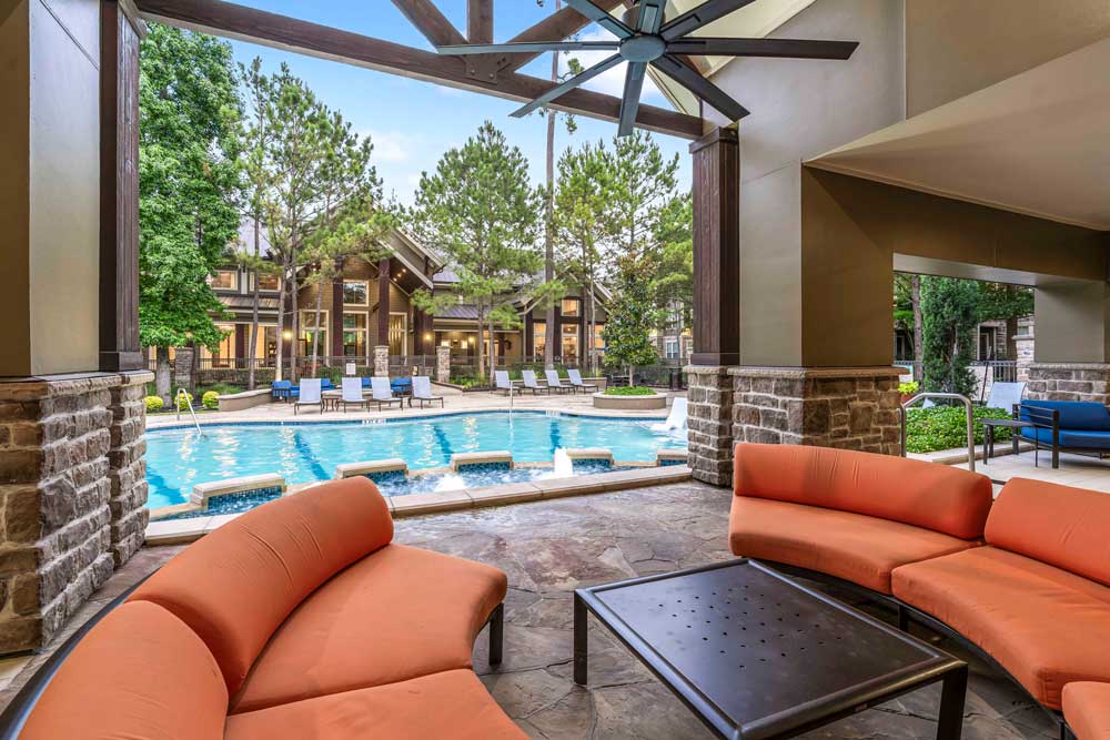 Poolside lounge with orange curved sofas, shaded ceiling fan, and a view of the pool and clubhouse beyond