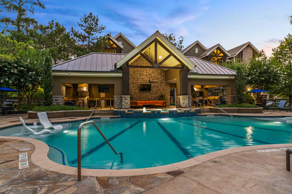 Pool area at dusk with soft lighting, built-in loungers, and clubhouse pavilion featuring a stone facade