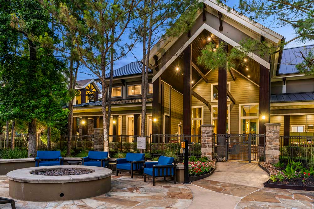 Clubhouse exterior at dusk with lit fire pit, blue outdoor seating, and pine trees in a cozy landscaped setting