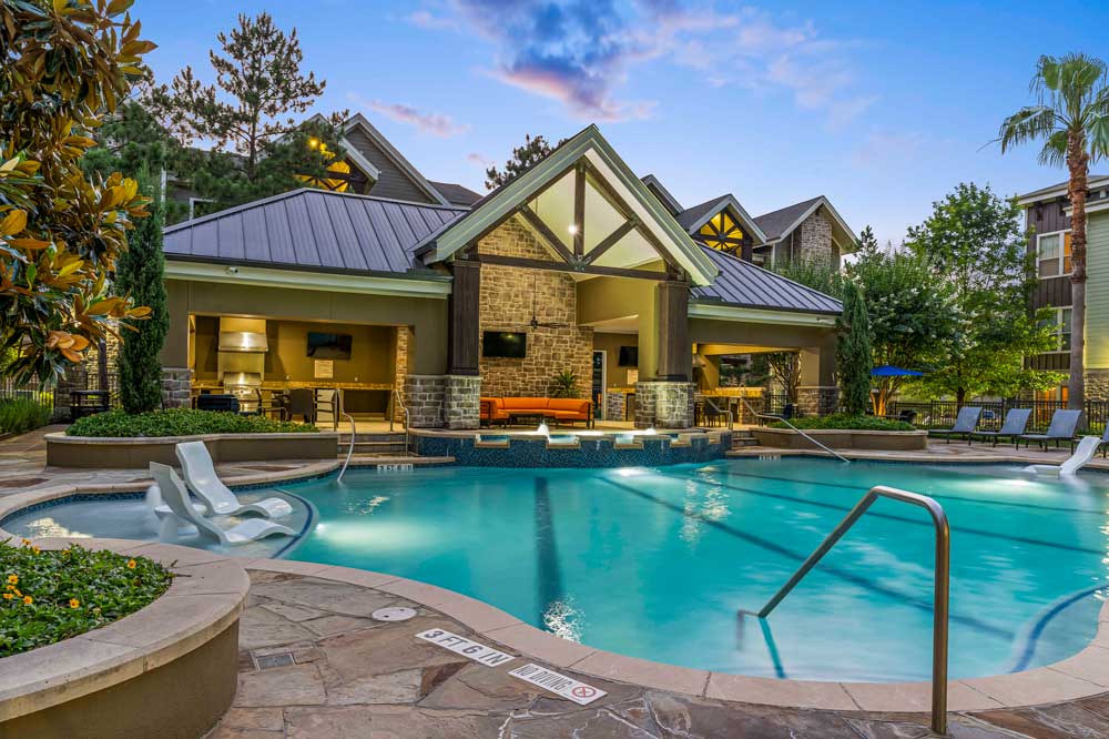 Resort-style pool at sunset with submerged loungers and a warmly lit clubhouse pavilion with stone accents